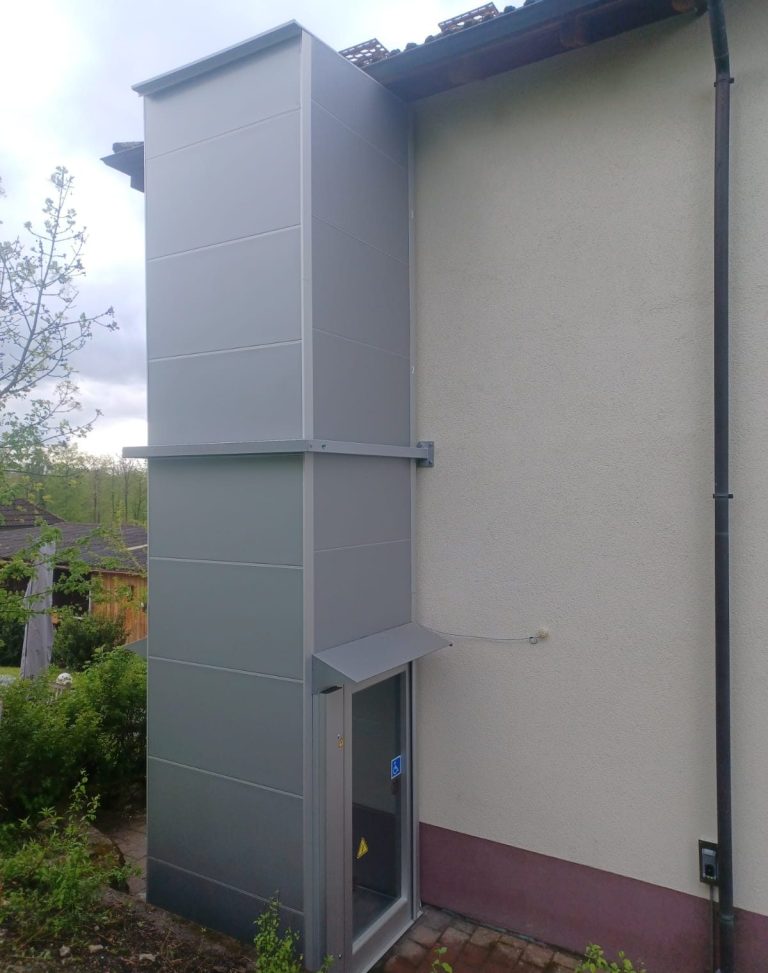 Außenansicht eines modernen Aufzugs im Gebäude, daneben Pflanzen und ein gepflegter Garten. Außenansicht eines modernen Aufzugs im Gebäude, daneben Pflanzen und ein gepflegter Garten.